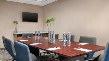 Un salón de conferencias con una larga mesa, sillas, botellas de agua y un televisor en la pared.