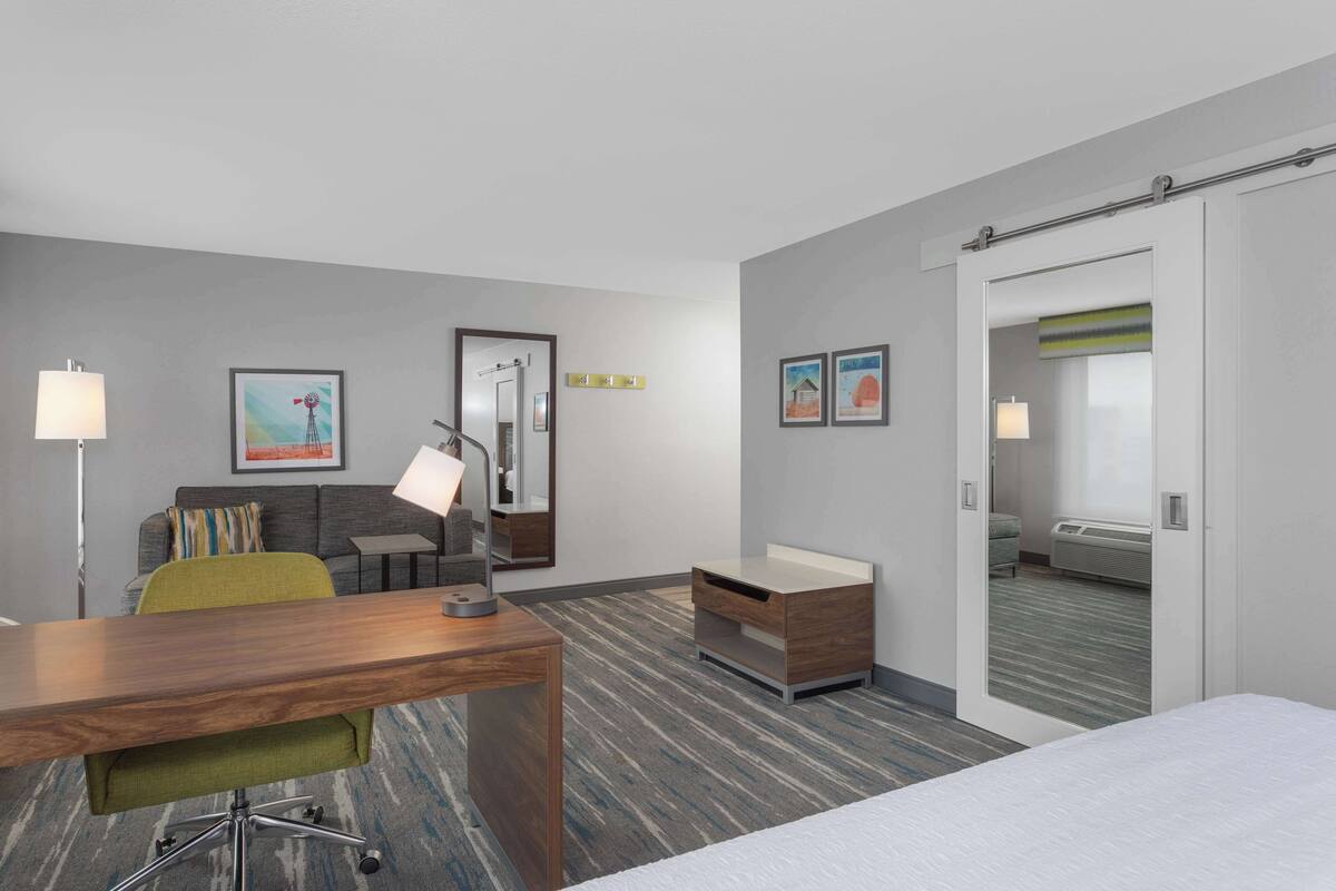 A hotel room with a wooden desk, green chair, a bed, a sofa, and a bathroom with a shower and toilet.