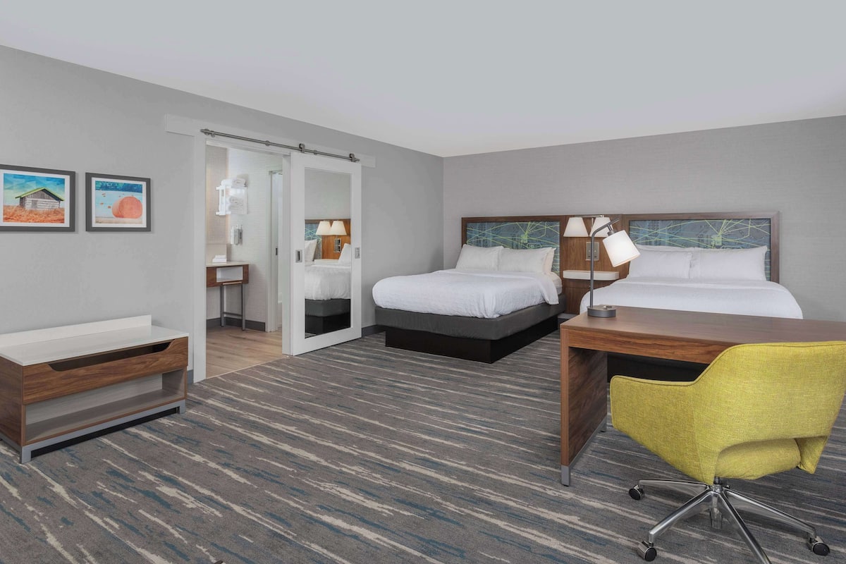 A hotel room with two beds, a desk, a chair, a bathroom with a shower, and a small table.