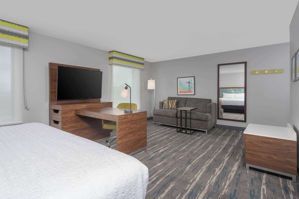 A hotel room with a large bed, a desk with a flat-screen TV, a sofa, a small table, and a mirror.