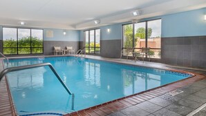 An indoor swimming pool with clear glass walls, a tiled floor, and a few chairs near the windows.