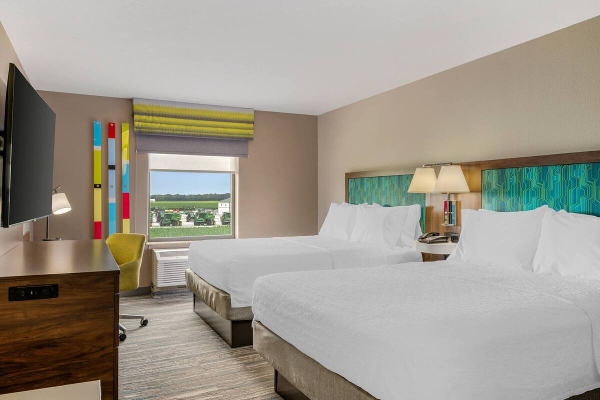 A hotel room with two beds, a desk, a TV, a window with a view, and colorful decor.