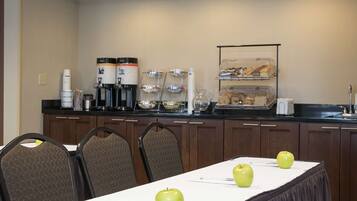 A breakfast bar with coffee makers, bowls, and a fruit display. There are chairs and a table with apples.