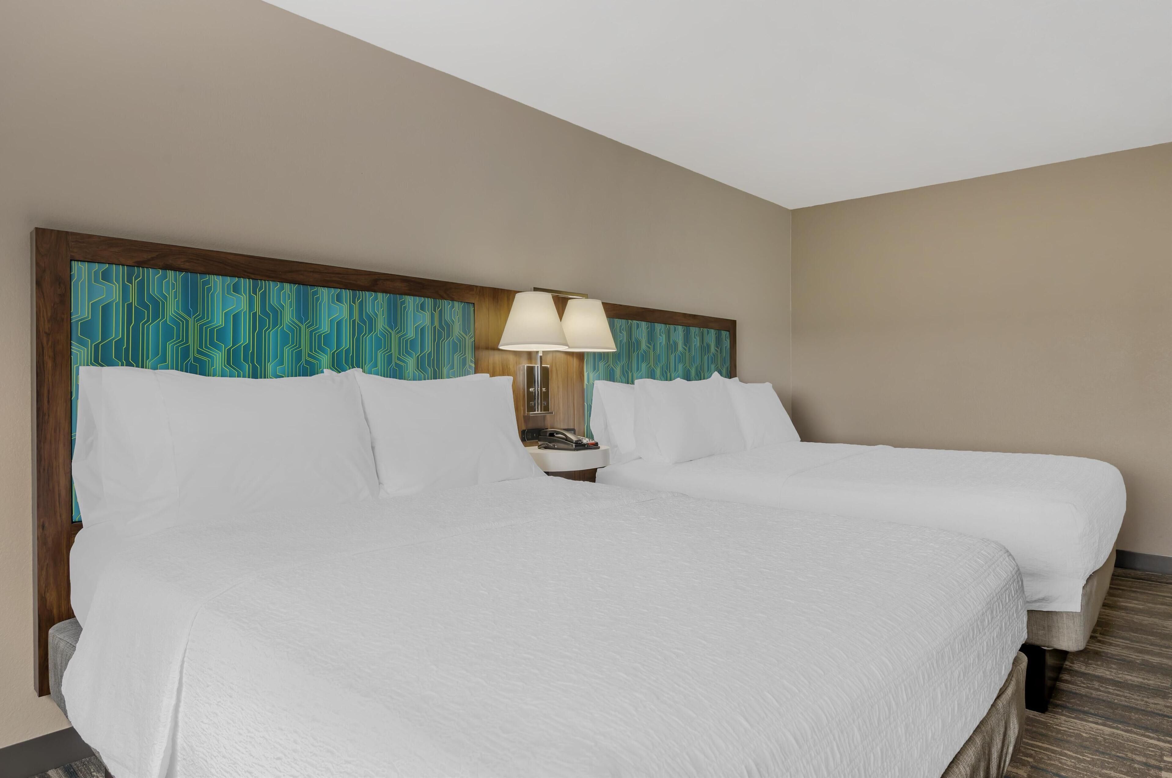 Ein Hotelzimmer mit zwei Betten, einem hölzernen Kopfende mit blauem Muster, einem Nachttisch mit Lampe und Blick ins Badezimmer.