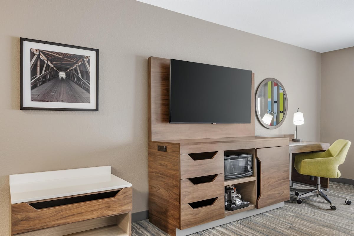 A hotel room with a wooden entertainment unit, a flat-screen TV, a desk with a chair, a framed picture, and a round mirror.