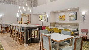 A modern dining area with multiple tables and chairs, a central island table, and artwork on the walls. - Hampton Inn & Suites Crawfordsville (Crawfordsville)