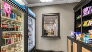 A hotel corridor with a beverage refrigerator stocked with various drinks, a mirror on the wall, and snack shelves.