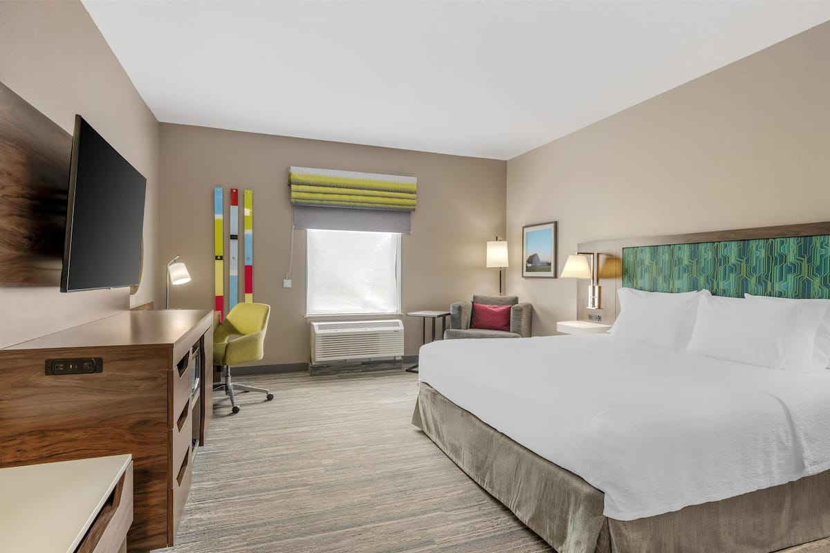 A hotel room with a large bed, a desk with a TV, a chair, a window with blinds, a lamp, and a painting on the wall.