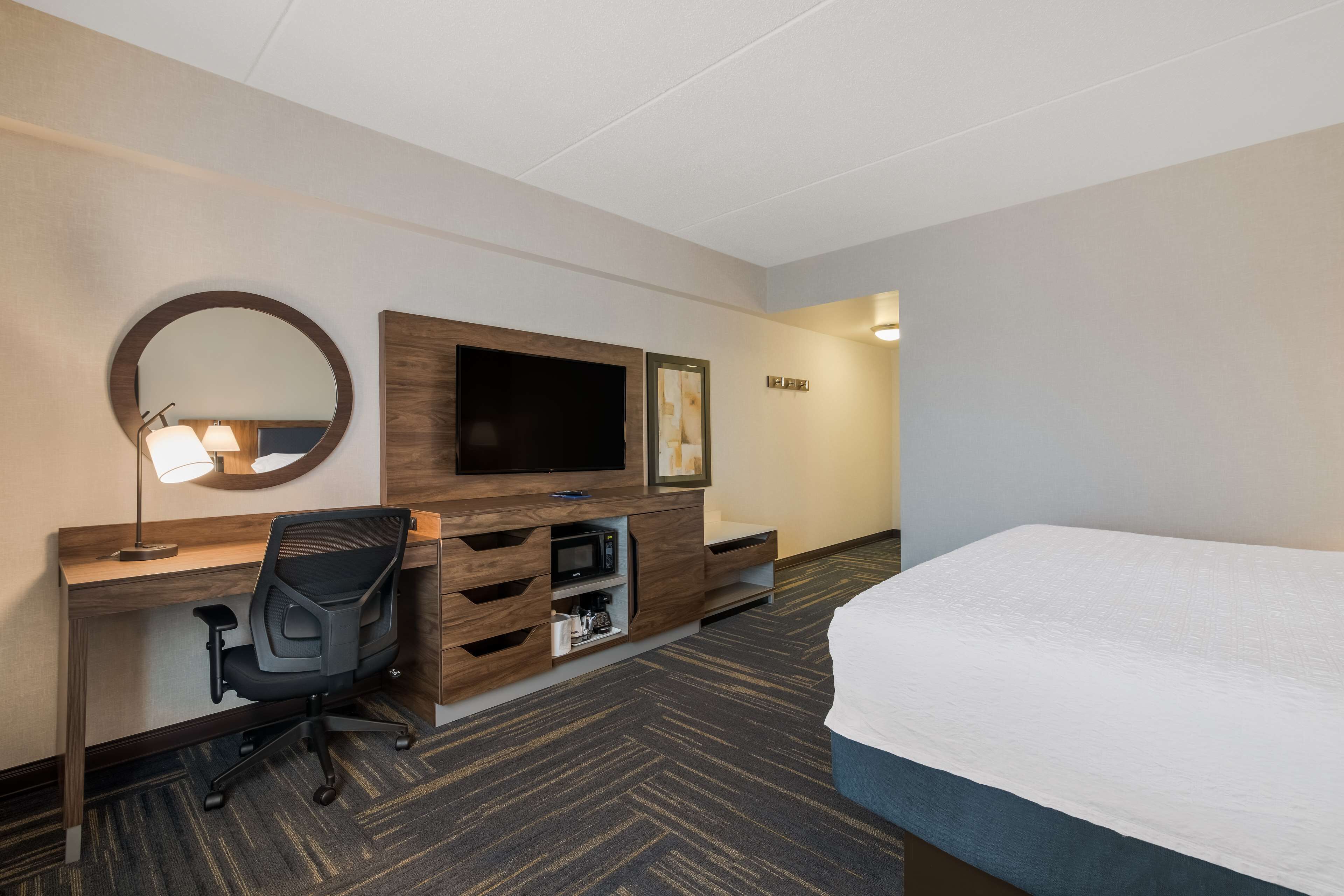 A hotel room with a bed, a desk with a chair, a television, a mirror, and a wall-mounted shelf.