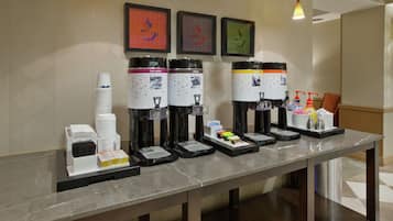 Una estación de bebidas de autoservicio con múltiples dispensadores, que incluyen agua caliente, café y té, y una bandeja con condimentos y servilletas.