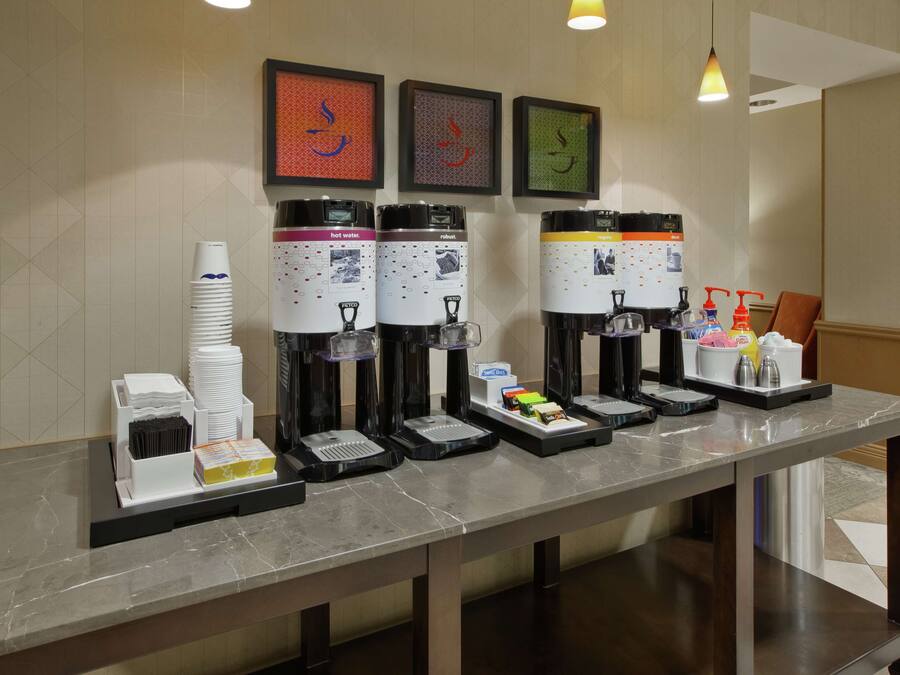 Una estación de bebidas de autoservicio con múltiples dispensadores, que incluyen agua caliente, café y té, y una bandeja con condimentos y servilletas.
