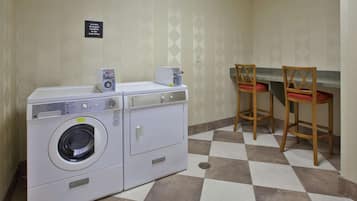 A laundry area with a washing machine, a dryer, and a sign indicating the location. There are two wooden chairs with red cushions.