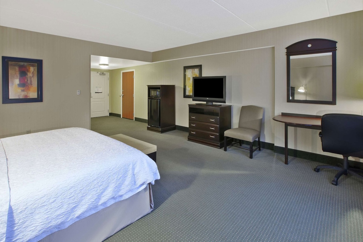 Quarto de hotel com cama, escrivaninha, cadeira, televisão e cômoda.