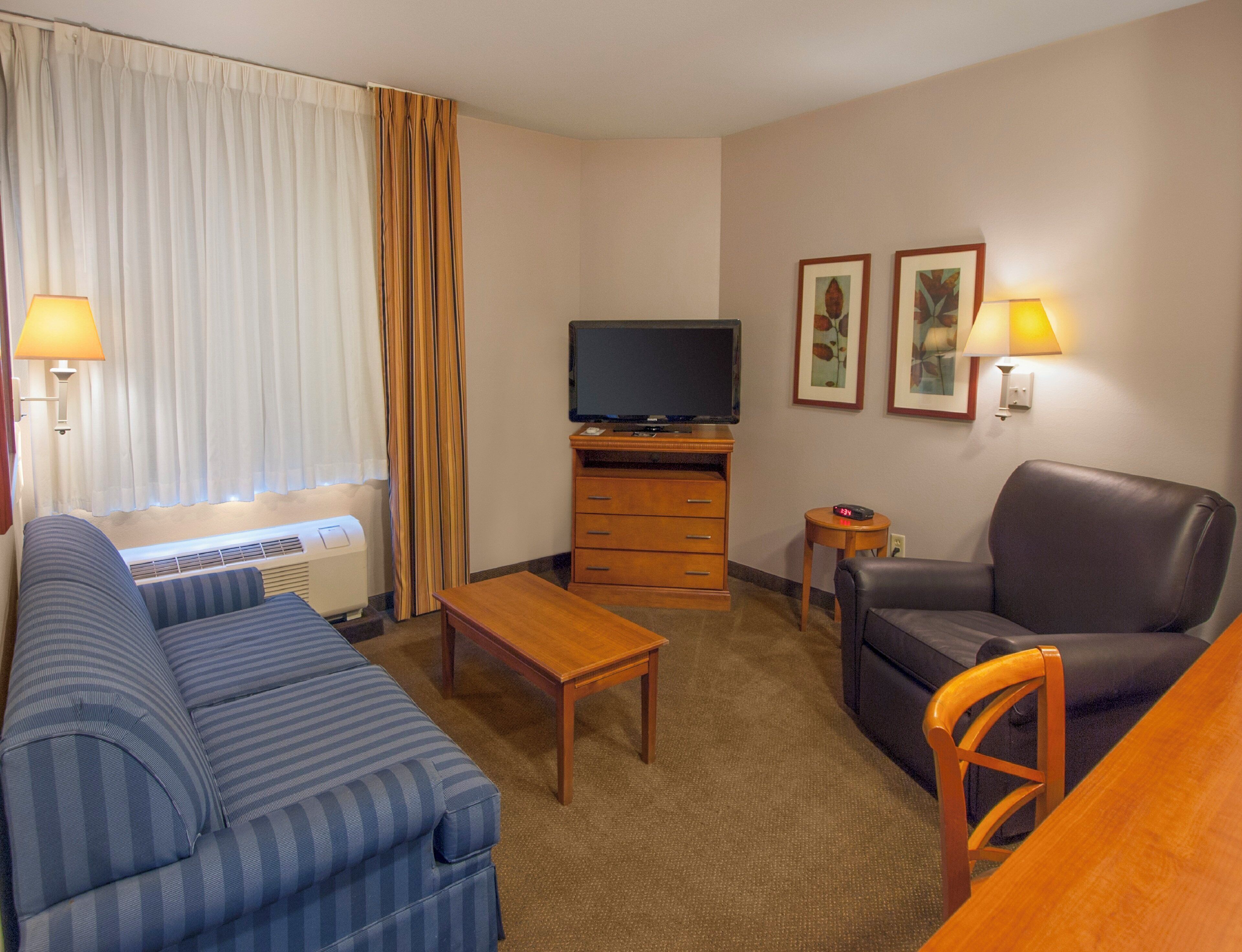 Habitación de hotel con sofá, televisor, mesa de centro, una silla y una mesita de noche con lámpara.