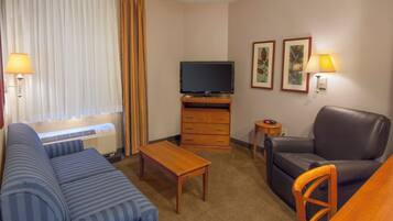 Habitación de hotel con sofá, televisor, mesa de centro, una silla y una mesita de noche con lámpara.