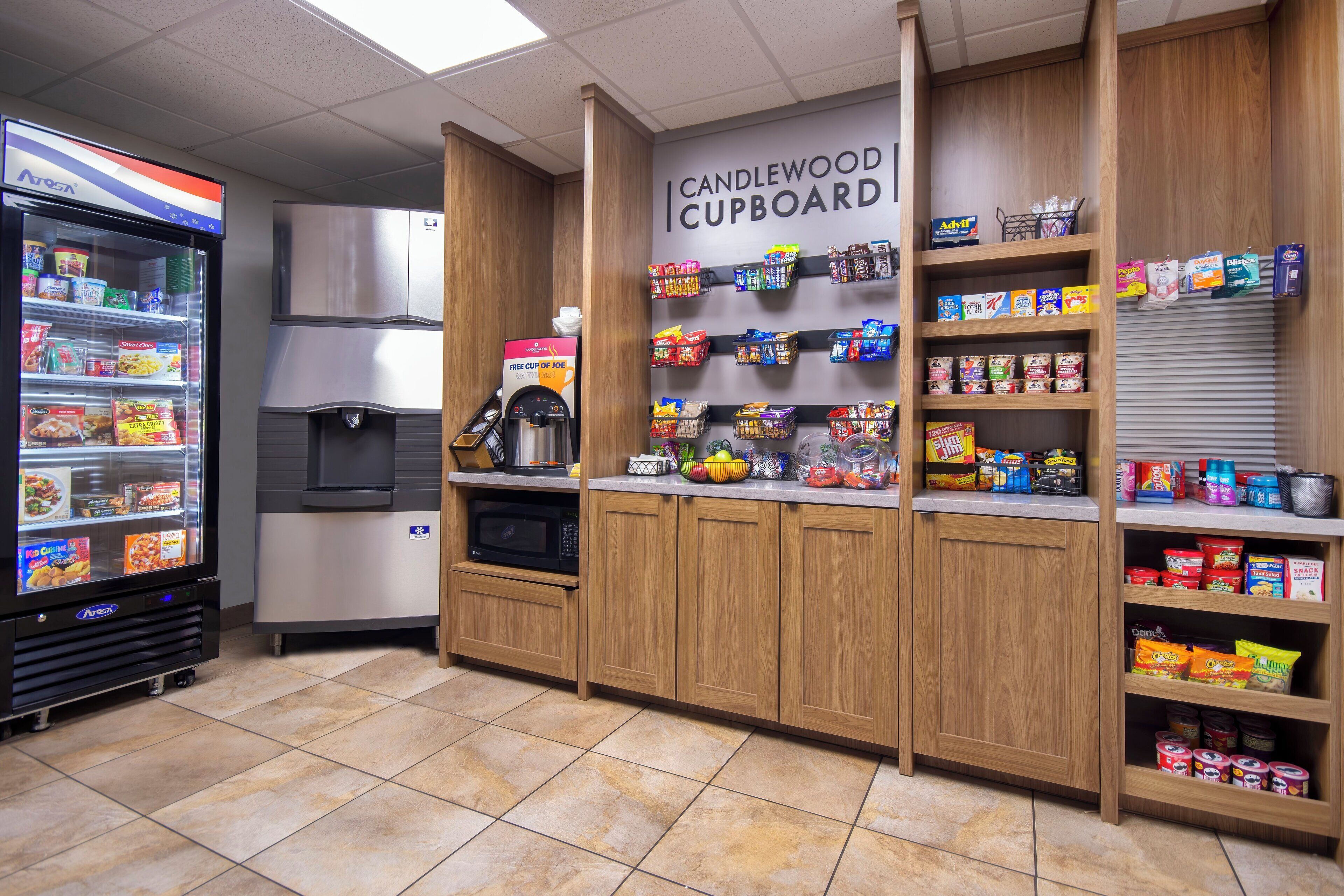 Una kitchenette con refrigerador, microondas y cafetera. Hay repisas con bocadillos y un letrero que dice "CANDLEWOOD CUPBOARD".