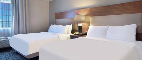 Habitación de hotel con dos camas, cabecero de madera, mesita de noche con lámpara y ventana con cortinas.
