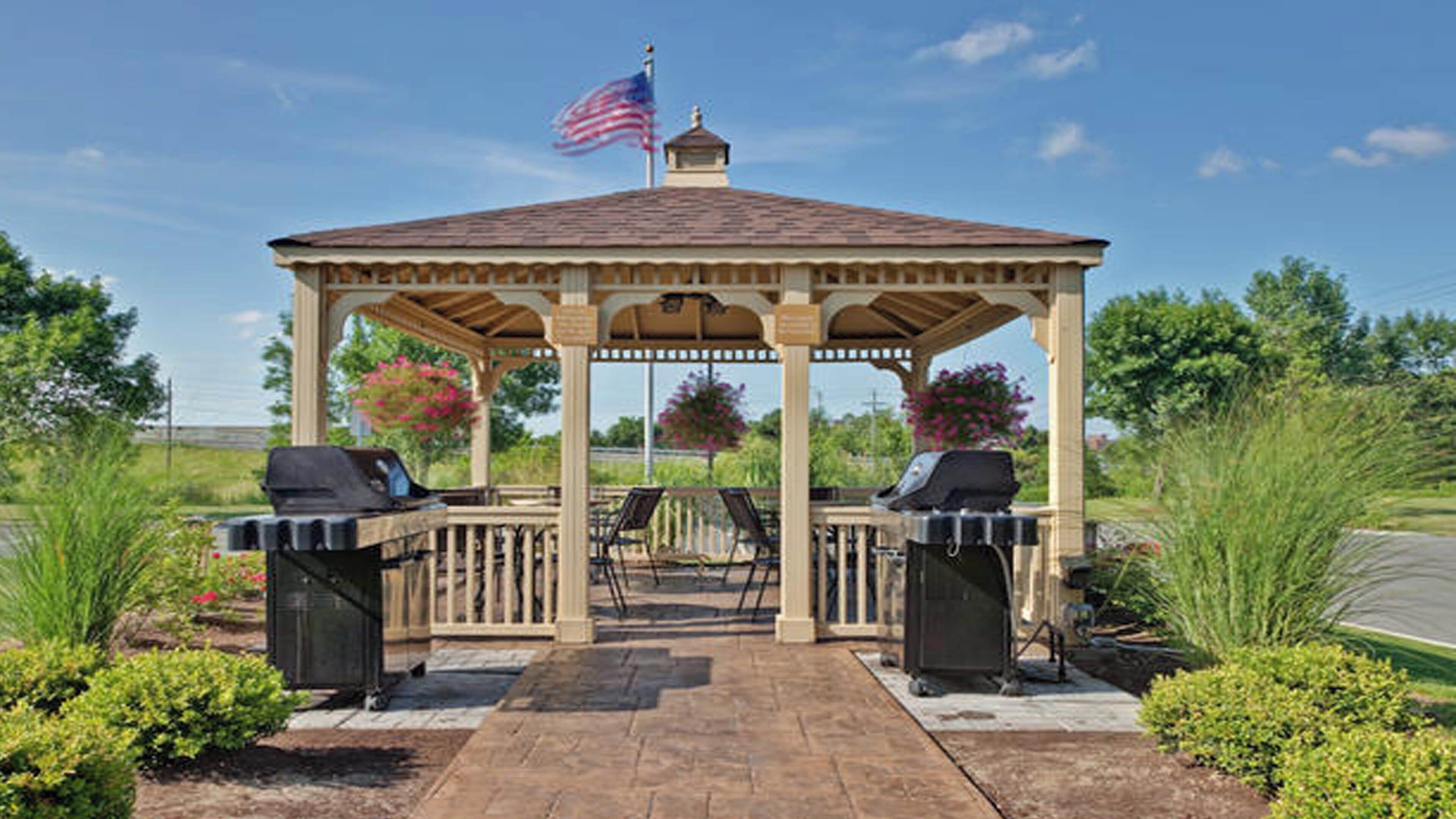 A gazebo with two barbecue grills, surrounded by landscaped gardens and a flag.