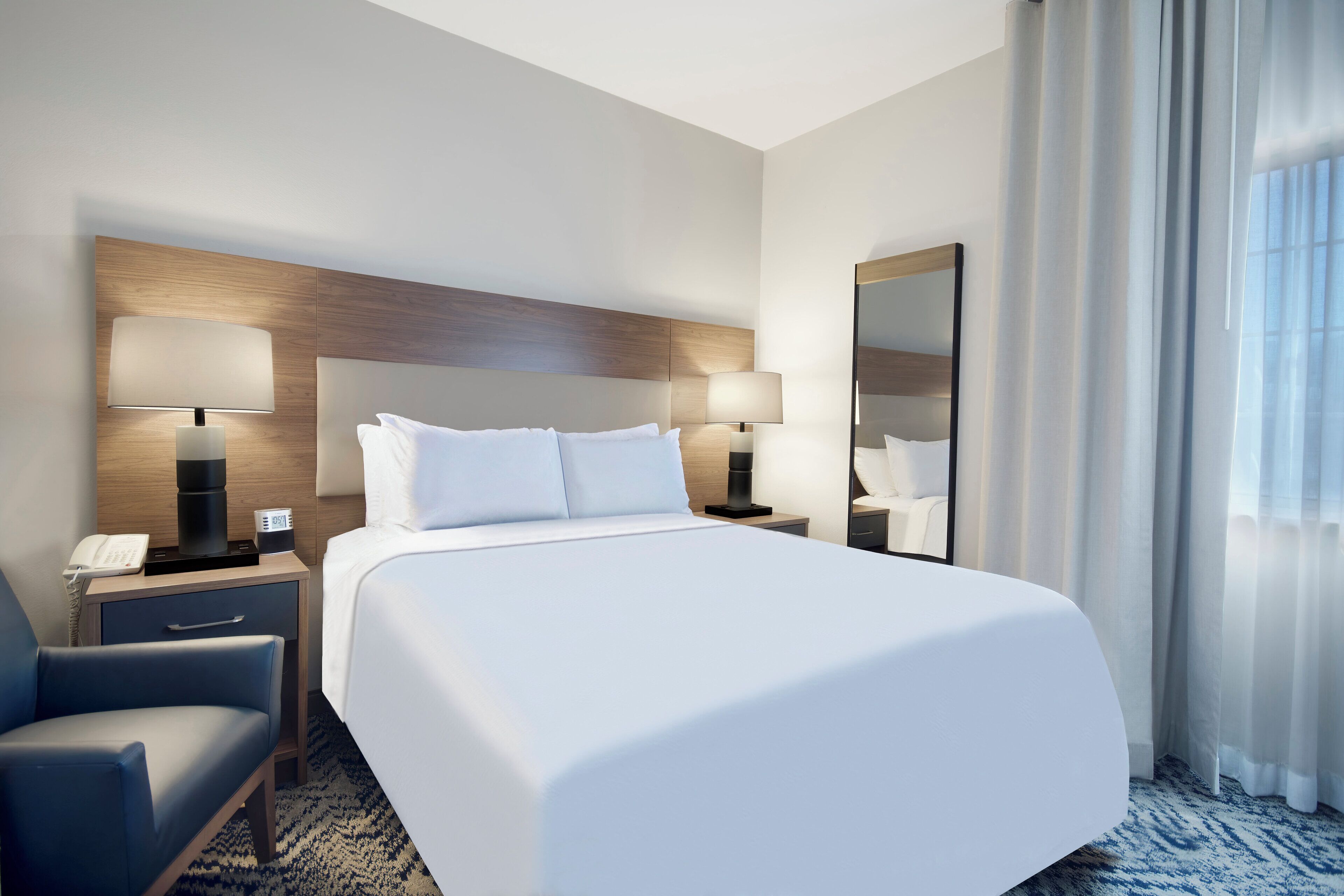 Et moderne hotelværelse med en stor seng, to sengeborde med lamper, en stol og et stort spejl.