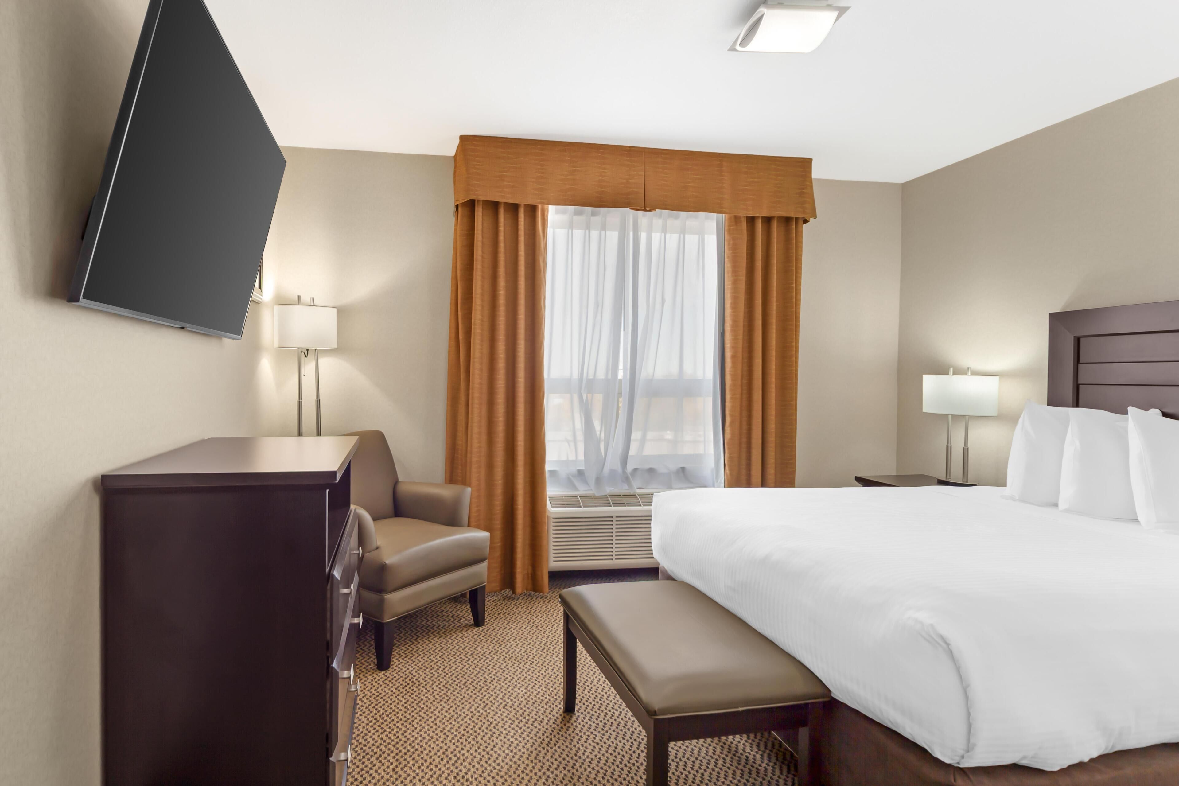 A hotel room with a bed, a desk, a chair, a TV mounted on the wall, and a window with curtains.