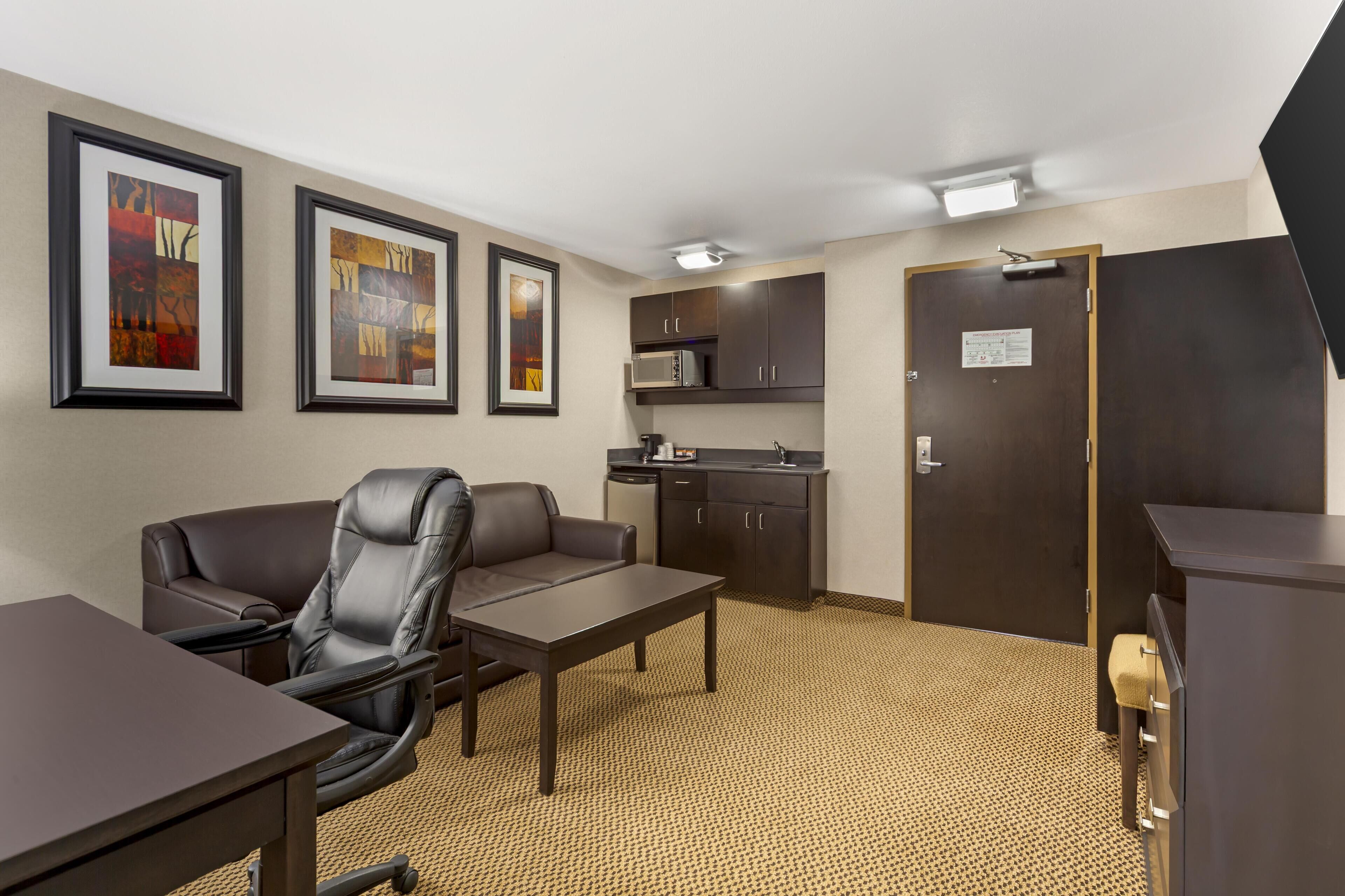 A hotel room with a desk, chair, kitchenette, and framed artwork on the wall.