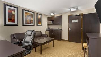A hotel room with a desk, chair, kitchenette, and framed artwork on the wall.