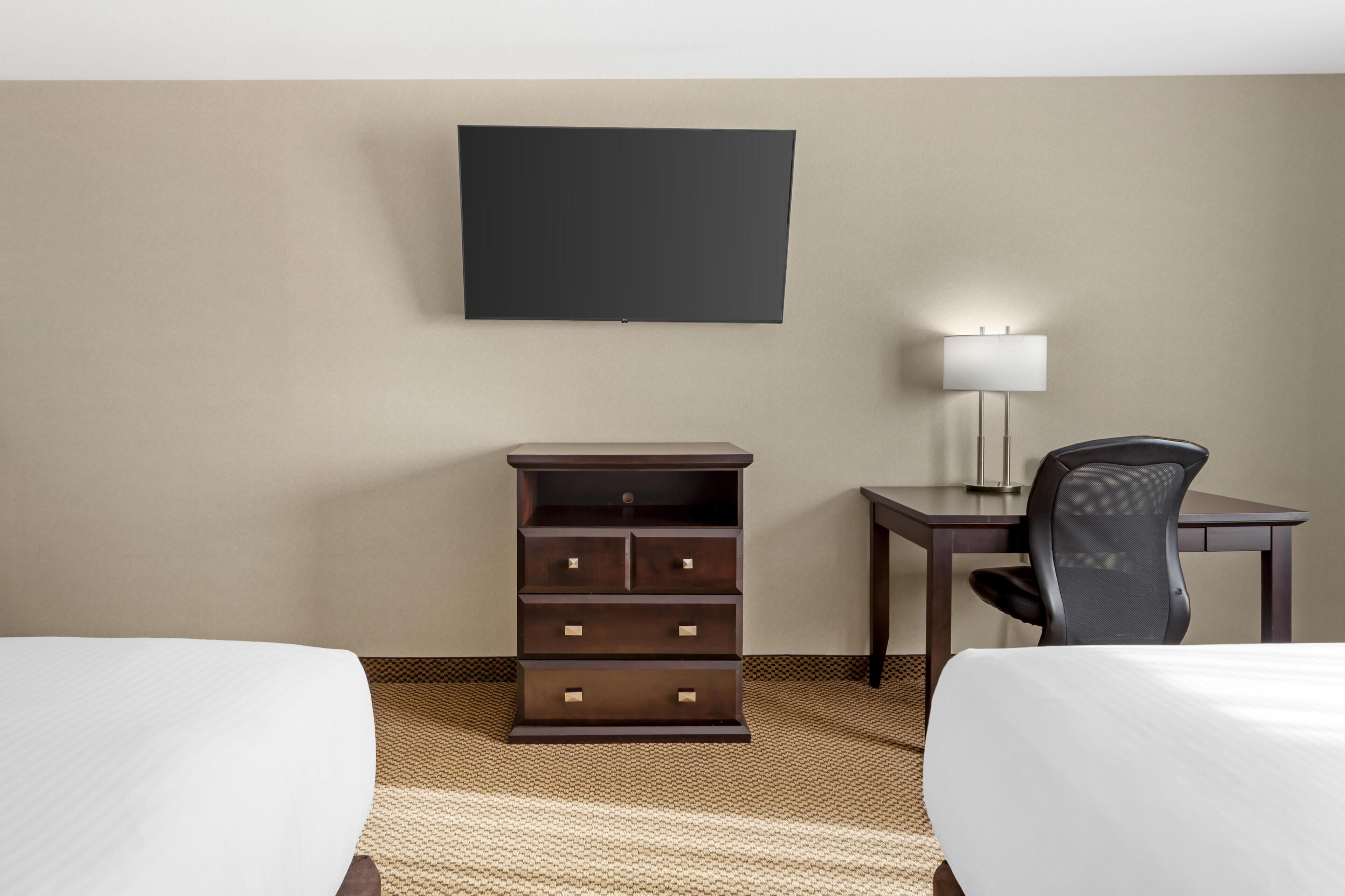 A hotel room with two beds, a desk, a chair, a TV, and a lamp.