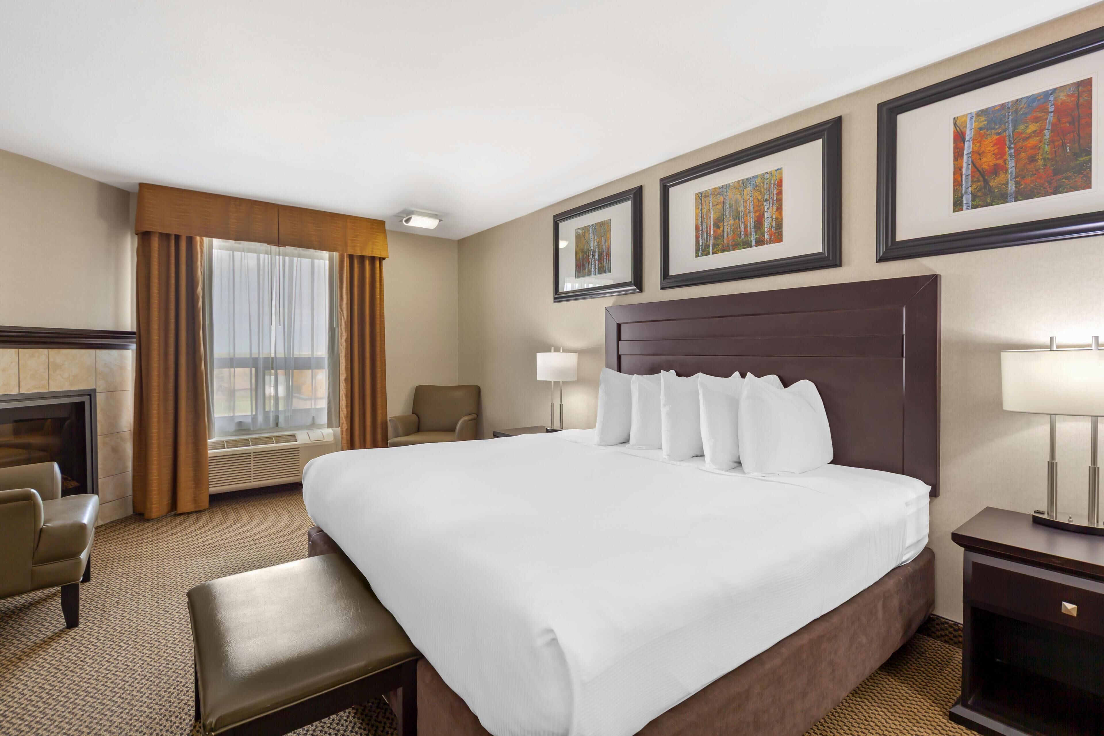 A hotel room with a large bed, a desk, a chair, a fireplace, and framed artwork on the wall.