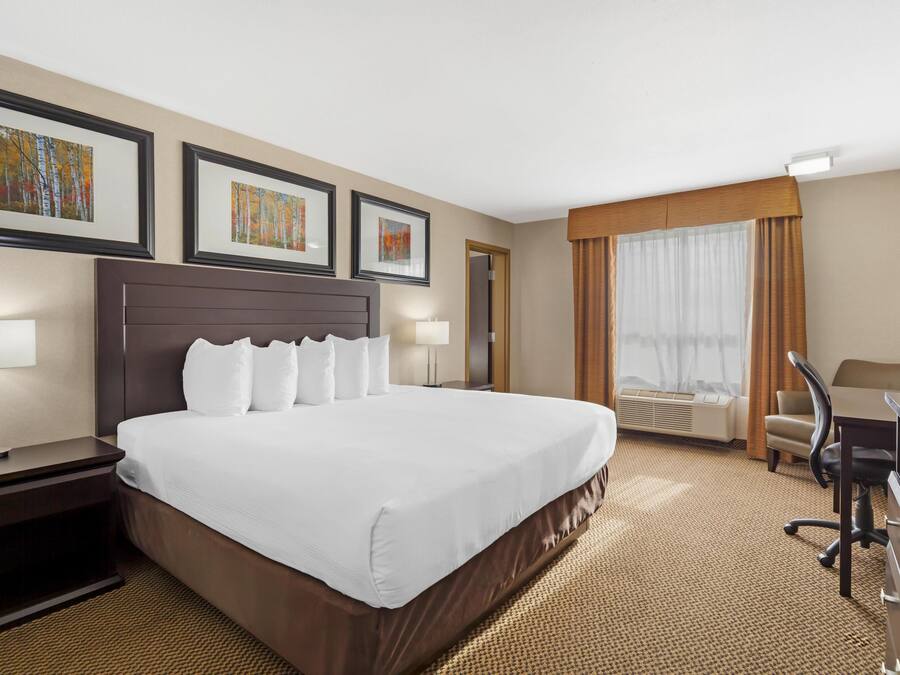 A hotel room with a large bed, a desk, and a chair. There are framed pictures on the wall, a window with curtains, and an air conditioning unit.