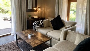 Un salon confortable avec un canapé, une table basse et une table d’appoint sur laquelle se trouve une lampe. Il y a une fenêtre avec des rideaux, un miroir et une vue sur la verdure à l’extérieur.