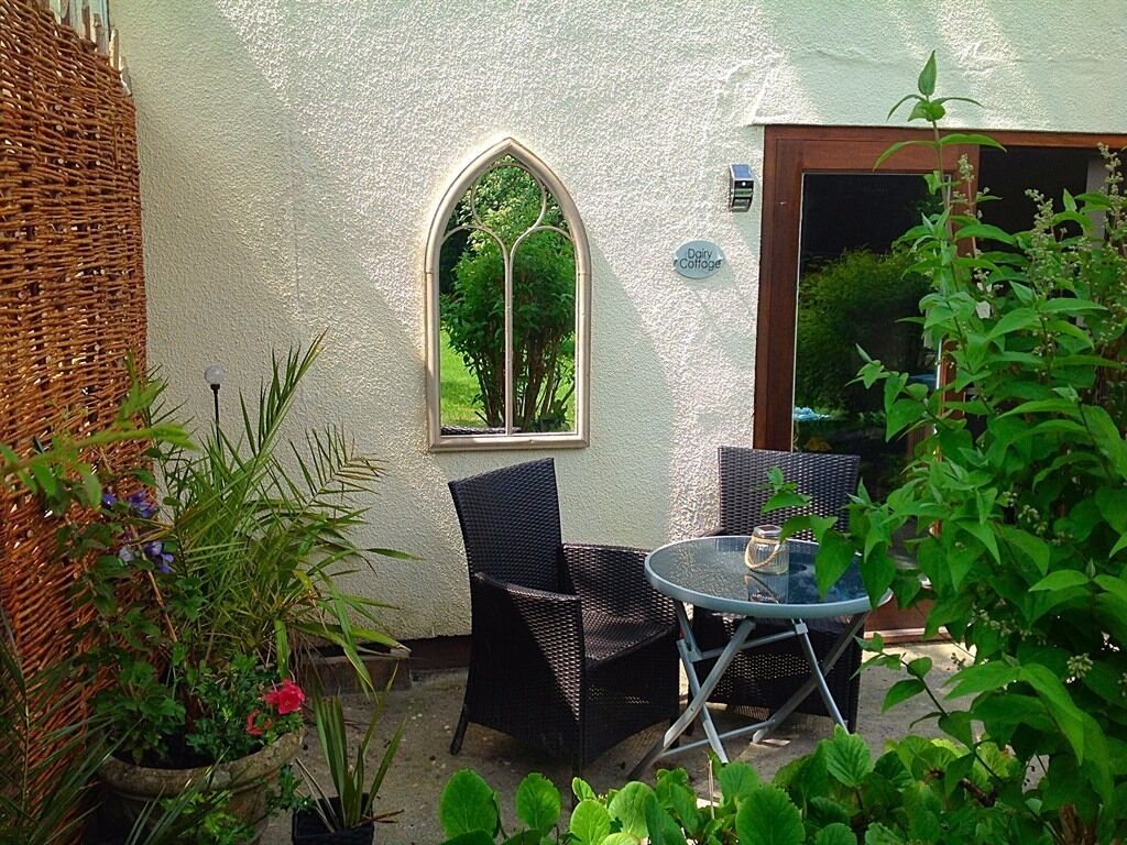 A small outdoor seating area with wicker chairs and a glass table, surrounded by potted plants and a white textured wall.