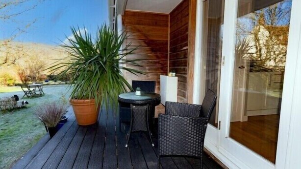 Une terrasse en bois avec une table et des chaises, une grande plante en pot et une porte-fenêtre coulissante donnant sur une pièce.