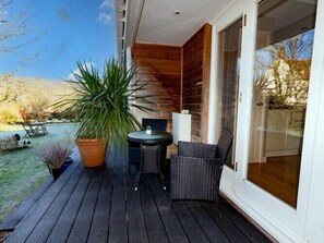 Una terraza de madera con una mesa y sillas, una planta grande en maceta y un ventanal corredera que da a una habitación.