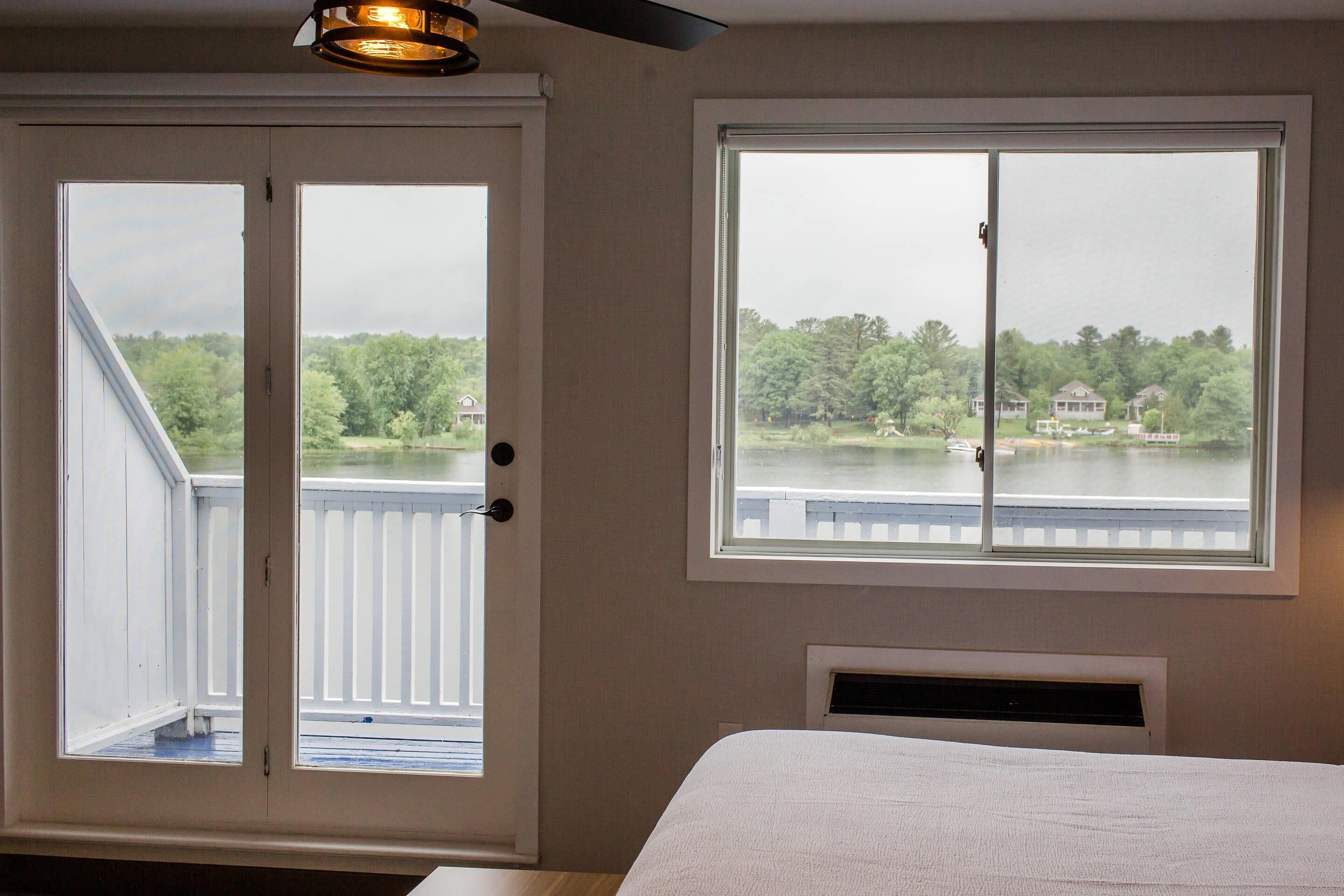 A bedroom with a large bed, a window with a view of a lake and houses, and a door leading to an outdoor balcony.