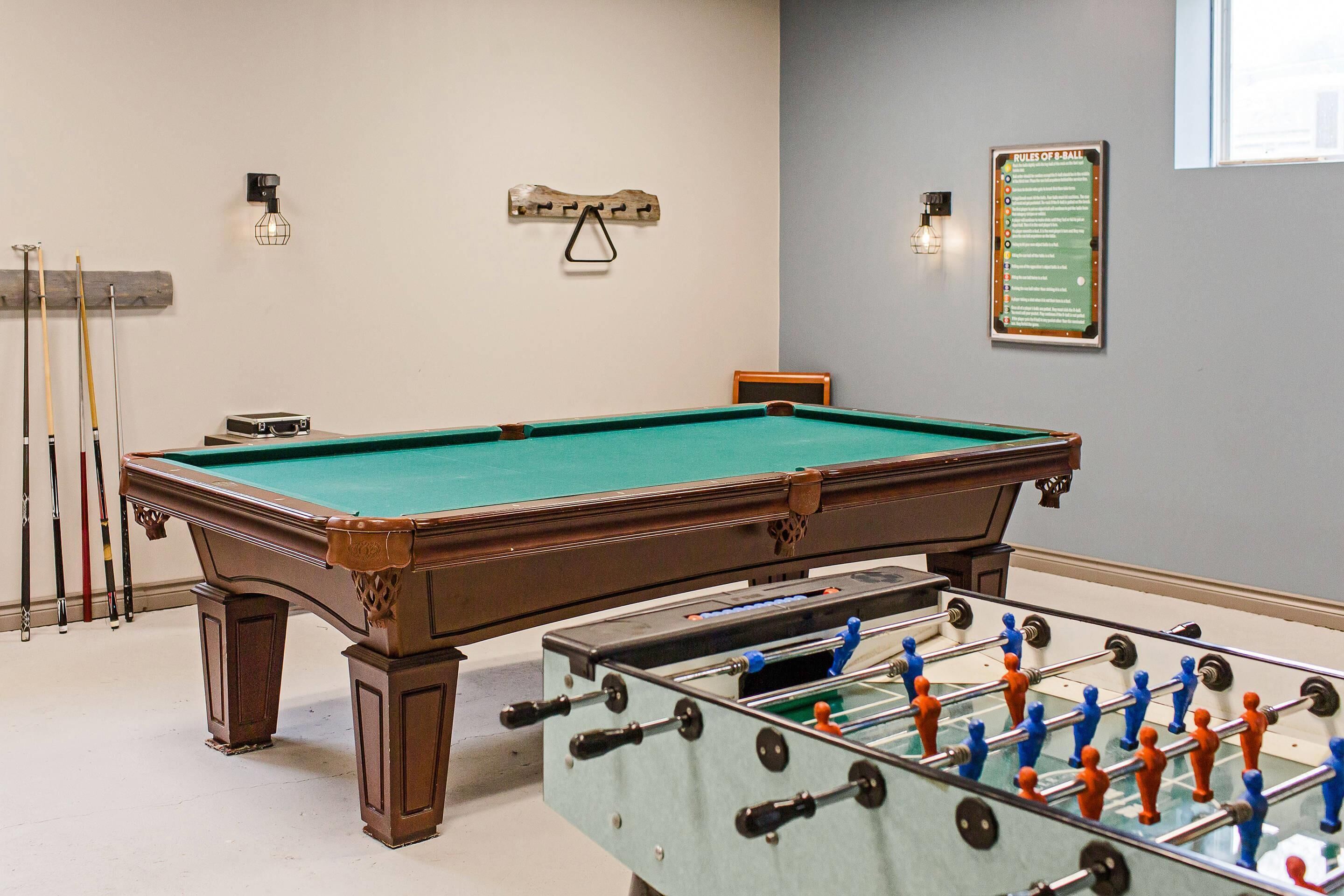 A pool table with a foosball table in front, a wall-mounted light fixture, and a rules of billiards sign.