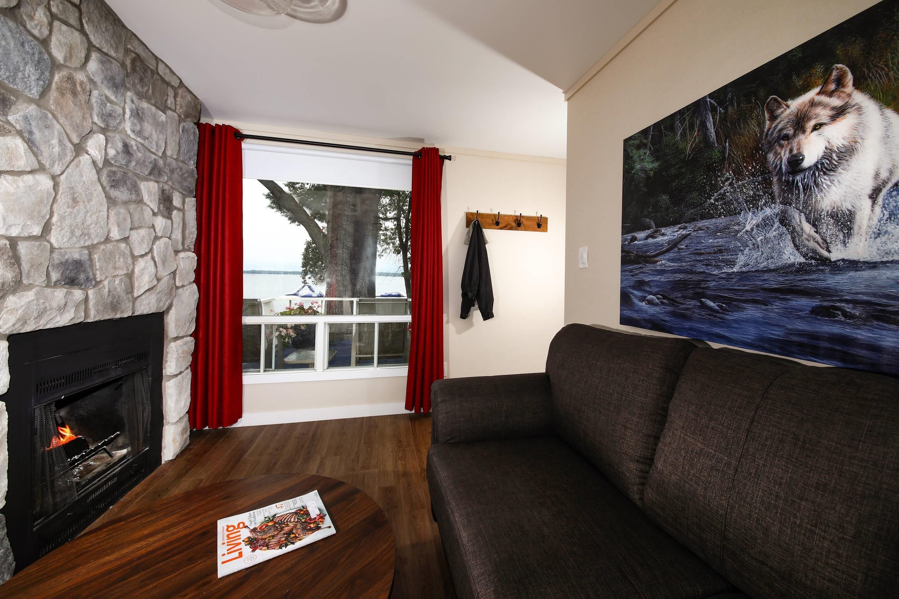 A living room with a stone fireplace, a large window with red curtains, a dark sofa, and a painting of a wolf.