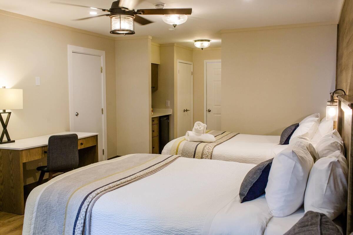 A hotel room with two beds, a desk, a chair, and a ceiling fan.