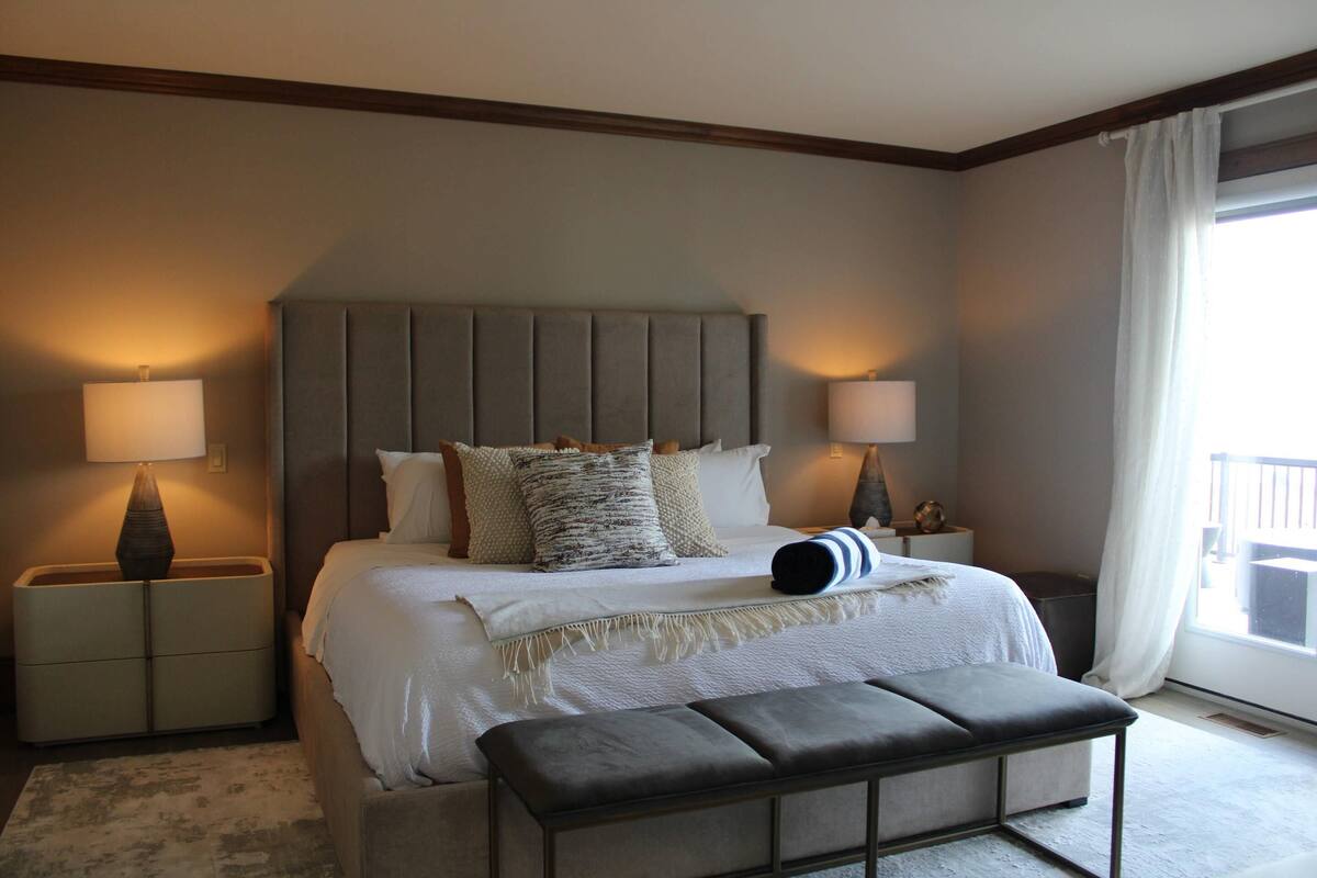 A bedroom with a large bed, two bedside tables, a bench, and a window with curtains.