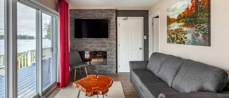 Ein Wohnzimmer mit grauem Sofa, einem Flachbildfernseher, einem Kamin aus Stein und einem großen Fenster mit Blick auf eine Terrasse.