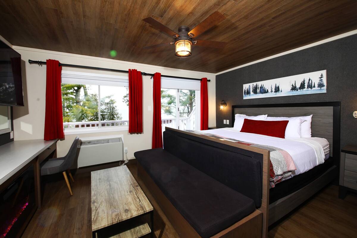 A bedroom with a bed, a desk, a chair, a sofa, a ceiling fan, and a window with red curtains.