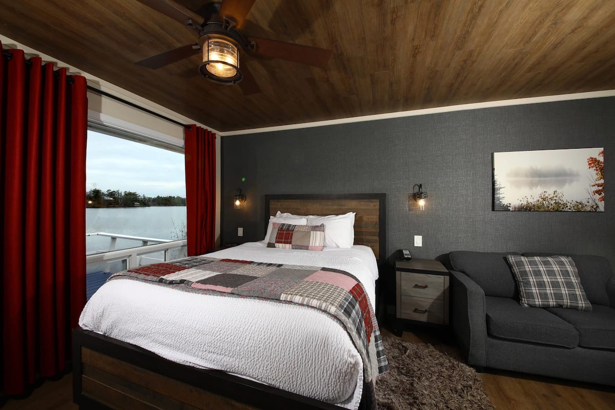 A bedroom with a bed, a nightstand, a sofa, a window with curtains, a ceiling fan, and a wall-mounted light fixture.