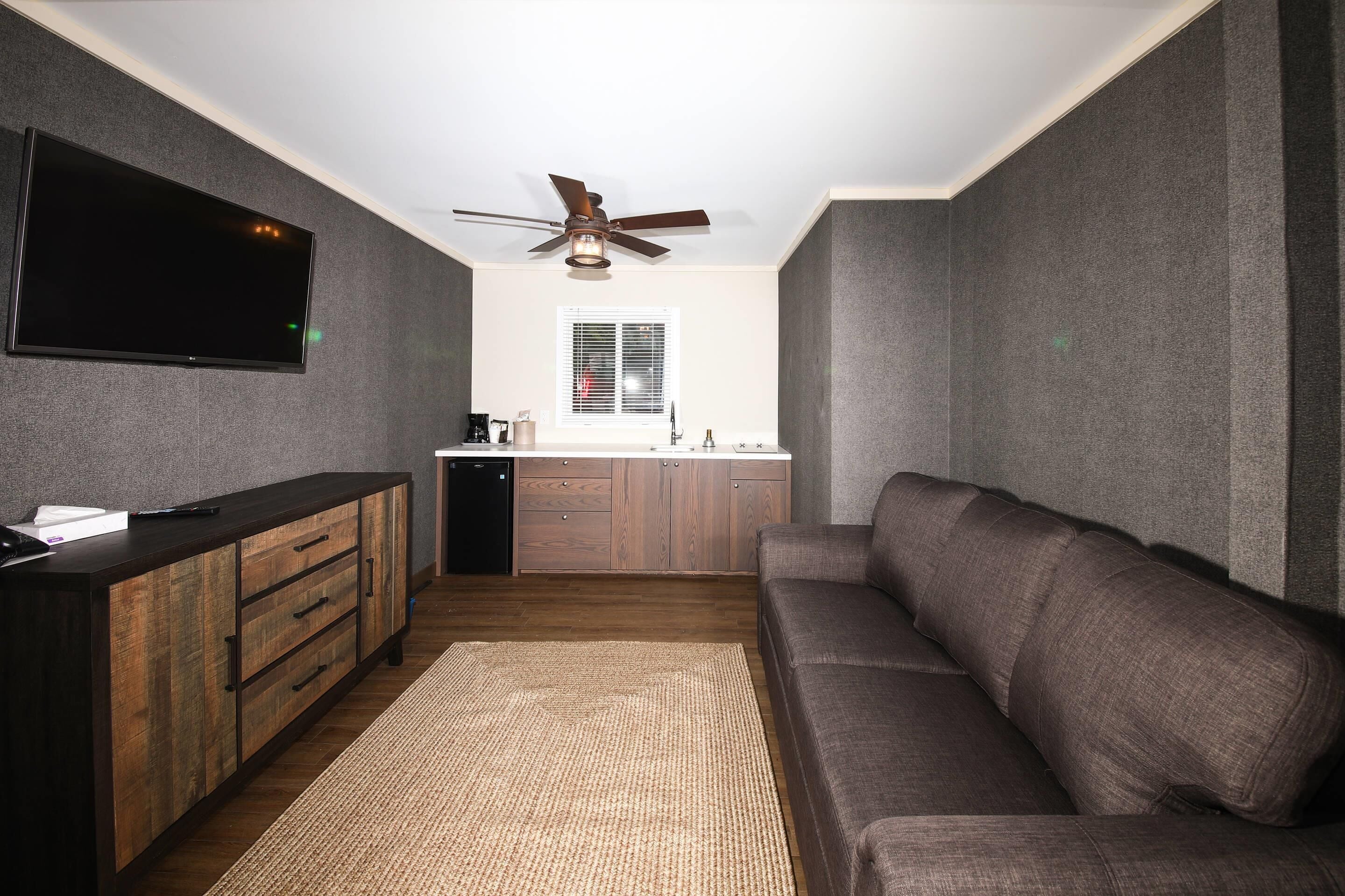 A compact living space with a kitchenette, a flat-screen TV mounted on the wall, a ceiling fan, and a sofa.