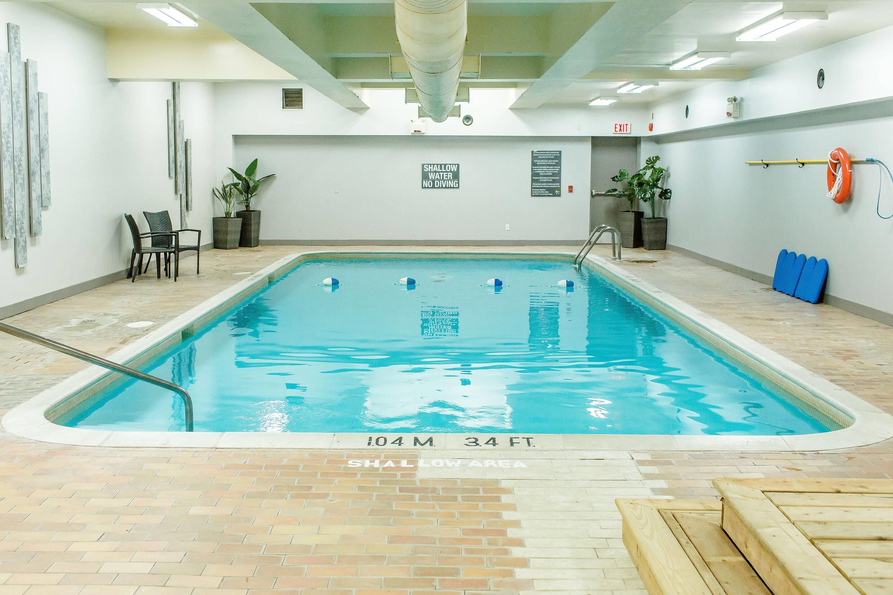 Piscina interior con señal de advertencia sobre poca profundidad, un flotador salvavidas y un cartel que indica las dimensiones de la piscina.