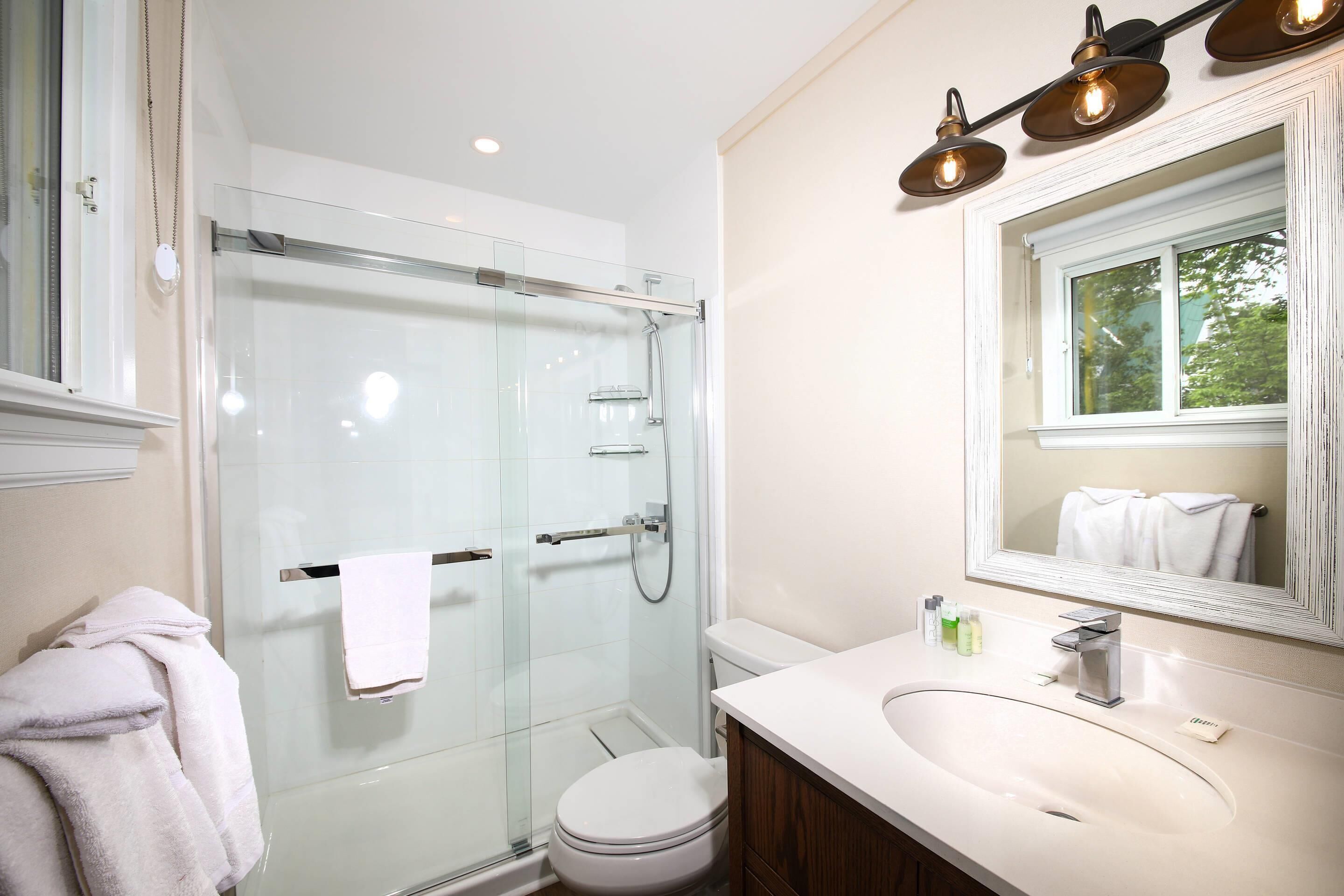 Un baño moderno con ducha envuelta en vidrio, un inodoro blanco, un lavamanos con espejo y una ventana con vistas a árboles.