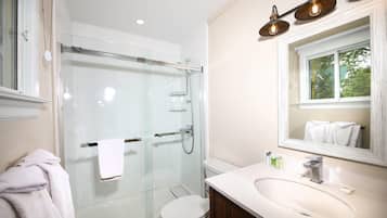 Un baño moderno con ducha envuelta en vidrio, un inodoro blanco, un lavamanos con espejo y una ventana con vistas a árboles.