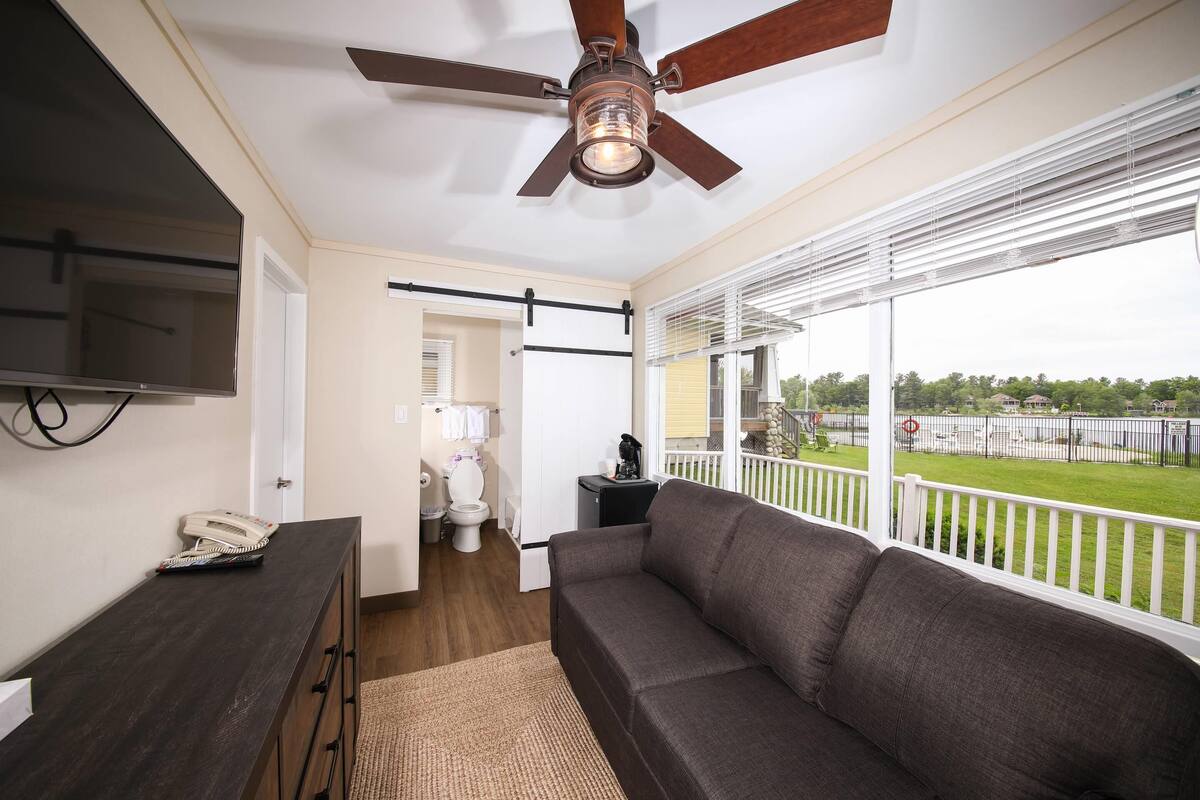 A modern living room with a dark brown sofa, a ceiling fan, a flat-screen TV mounted on the wall, and a large window offering a view of a green landscape.