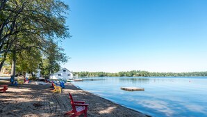 Ein Strand am See mit einem weißen Gebäude, roten und gelben Liegestühlen und einer anlegenden Mole.