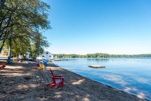 Une plage au bord d’un lac, avec un bâtiment blanc, des chaises de plage rouges et jaunes, et une jetée qui s’avance dans l’eau.