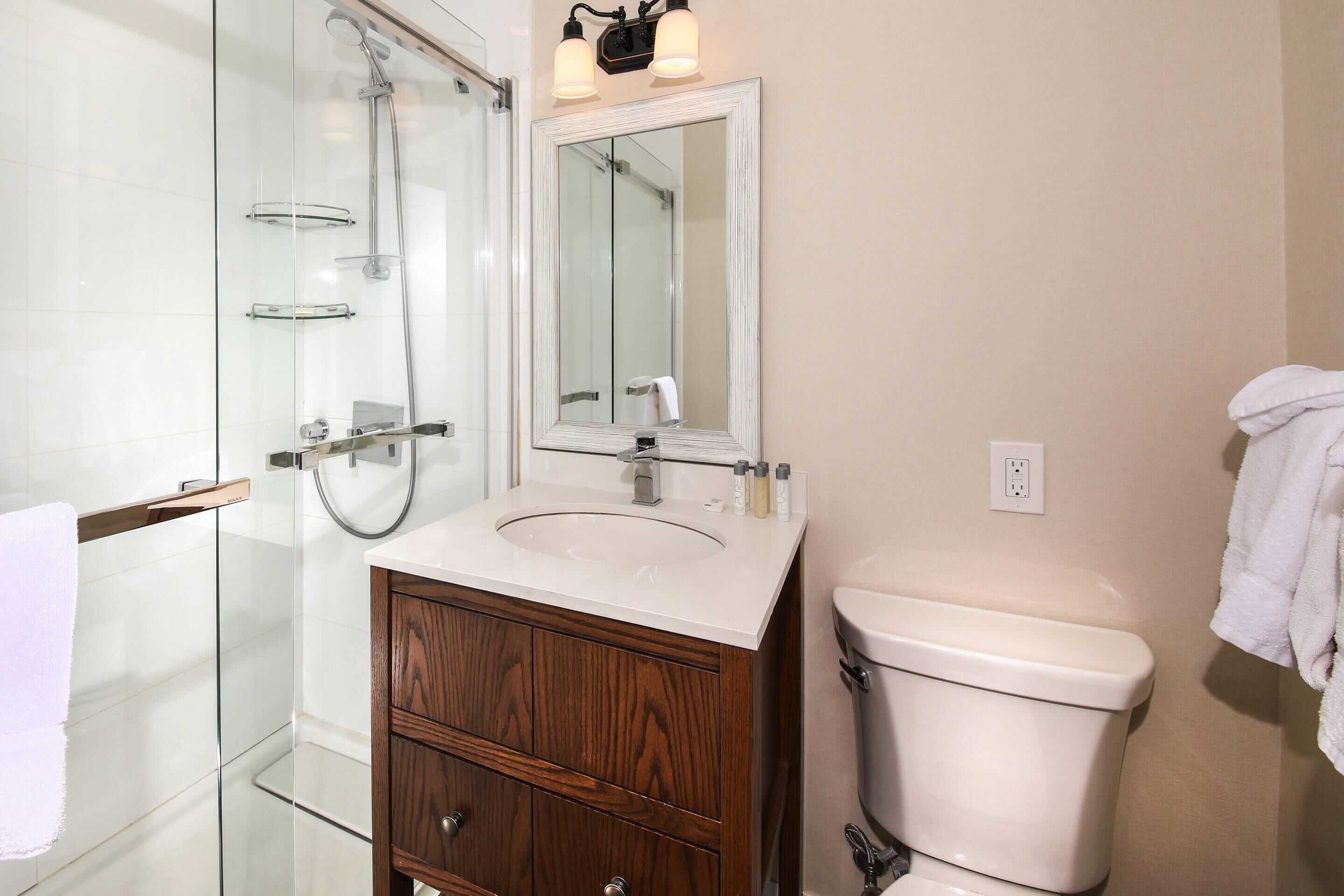 A bathroom with a glass shower enclosure, a white sink with a wooden vanity, a toilet, and a mirror above the sink.