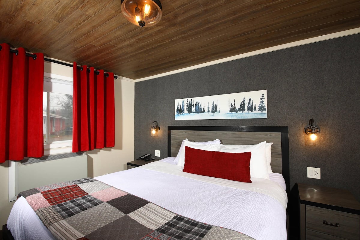 A neatly made bed with a quilt, a red pillow, and a headboard. There is a painting of trees on the wall and a window with red curtains.